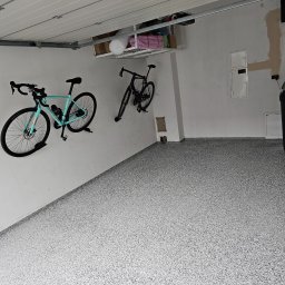JWK Sp. z o.o. - Gładka, jasnoszara posadzka żywiczna w garażu z widocznymi rowerami przymocowanymi do ściany i metalową półką pod sufitem.