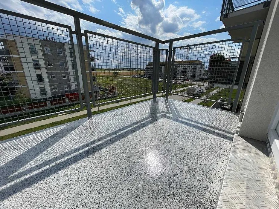 Szary balkon z metalową kratą i podłogą z żywicy epoksydowej w jasnych odcieniach szarości, z widokiem na budynki mieszkalne i zielony krajobraz.