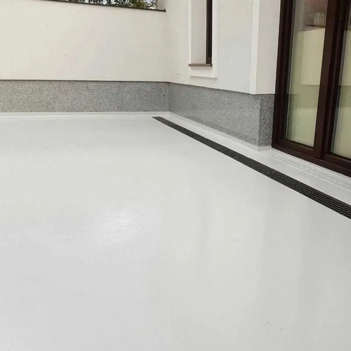 Biała, gładka posadzka z odpływem liniowym przy ścianie wykończonej szarym kamieniem dekoracyjnym, widoczne przesuwne drzwi balkonowe z brązowymi ramami.