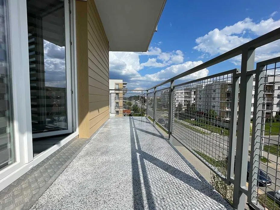 Wykończony balkon z szarą, nakrapianą posadzką i metalową balustradą z siatki, widok na osiedle mieszkaniowe w słoneczny dzień.
