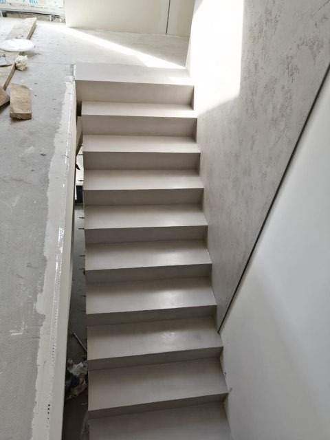 Schody wewnętrzne wykończone szarym mikrocementem, widok z góry na dół, z widocznymi śladami prac wykończeniowych na ścianach i podłodze.