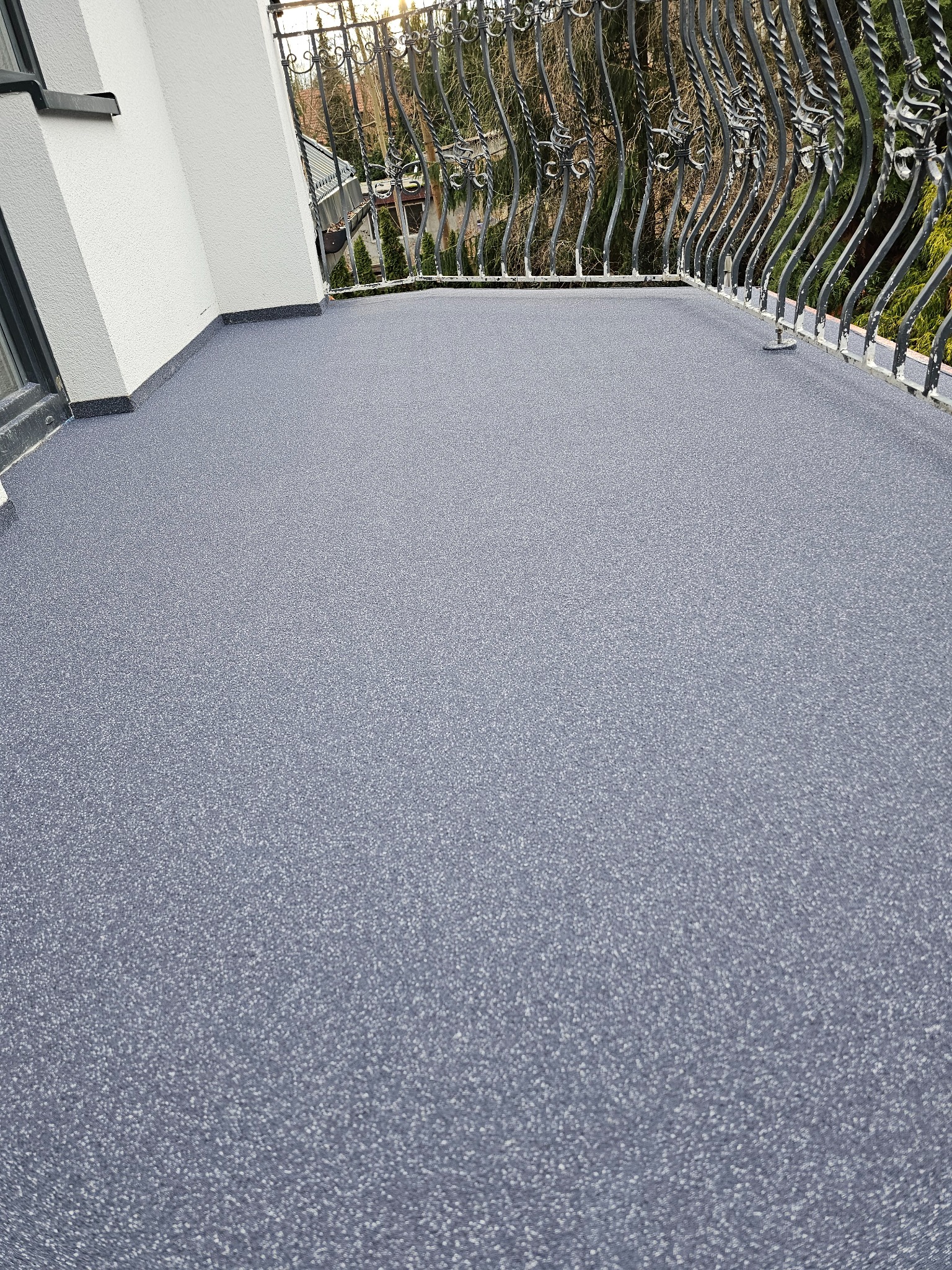 Nowoczesny balkon z szarą, drobnoziarnistą posadzką, wykończony kutą, metalową balustradą z dekoracyjnymi elementami, z widokiem na drzewa w tle.