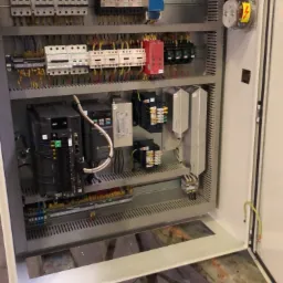 Otwarta szafa sterownicza z widocznymi zabezpieczeniami, stycznikami, sterownikami PLC i okablowaniem wewnątrz, zamontowana na metalowej konstrukcji.