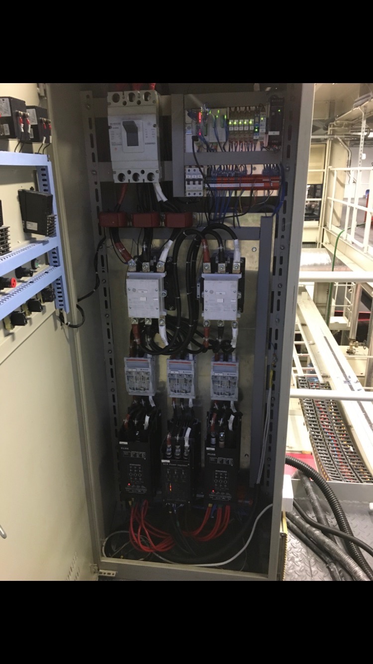 Otwarta szafa sterownicza z widocznymi stycznikami, bezpiecznikami, listwami zaciskowymi i przewodami, oświetlona jarzeniówką, z elementami infrastruktury w tle.