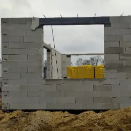 Wznoszenie ścian domu z betonu komórkowego Ytong, widoczny niezabezpieczony wykop fundamentów i paleta żółtych bloczków izolacyjnych w otworze okiennym.