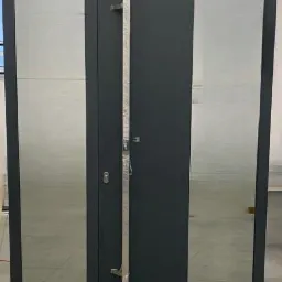 Nowoczesne, antracytowe drzwi wejściowe z dwoma panelami szklanymi, zabezpieczone folią ochronną na metalowych elementach, stojące na betonowej posadzce w jasnym wnętrzu.