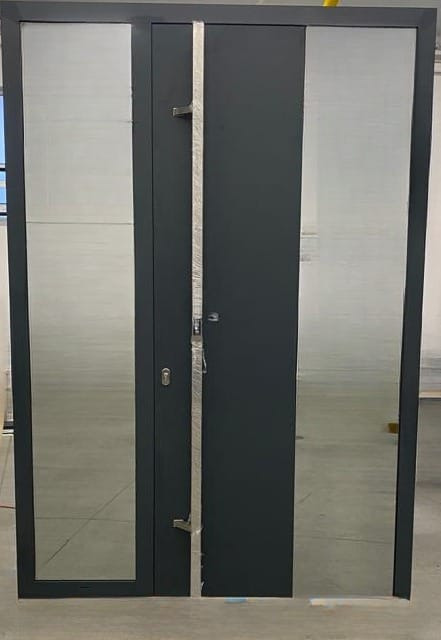 Nowoczesne, antracytowe drzwi wejściowe z dwoma panelami szklanymi, zabezpieczone folią ochronną na metalowych elementach, stojące na betonowej posadzce w jasnym wnętrzu.