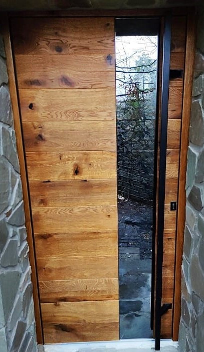Drewniane drzwi wejściowe z poziomych desek, z czarną metalową ramą i przeszkleniem, widok na zewnątrz, kamienna elewacja budynku.