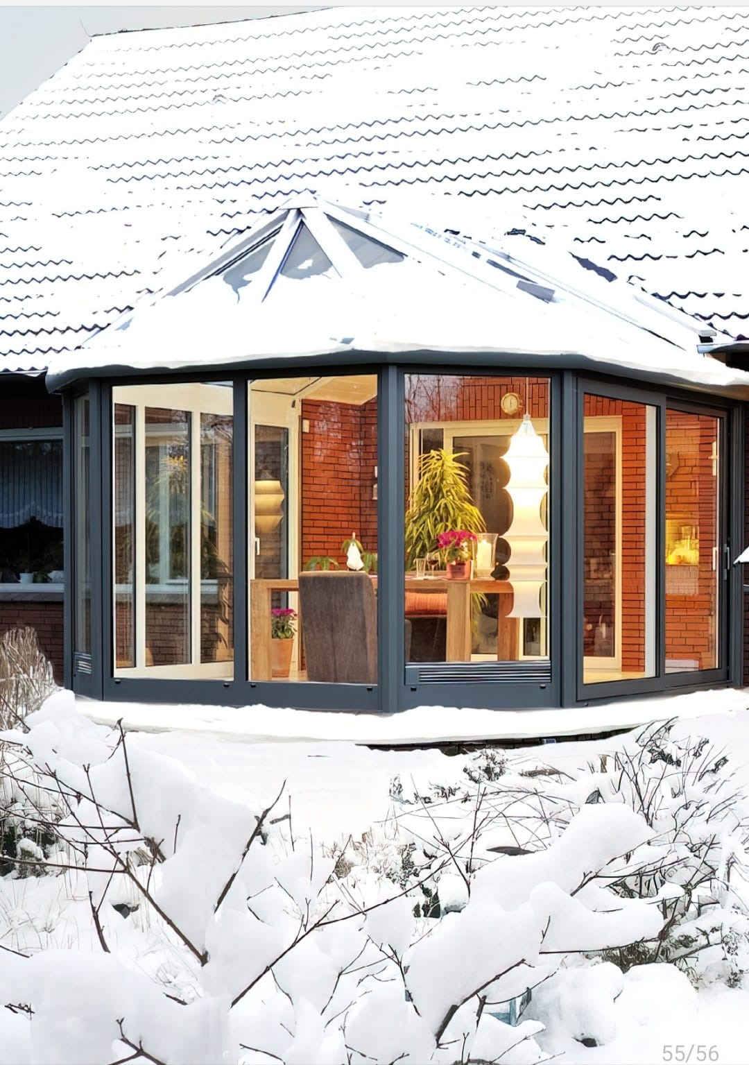 Zimowy ogród zimowy z szarymi ramami okiennymi, czerwona ceglana ściana i śnieżny krajobraz na pierwszym planie.