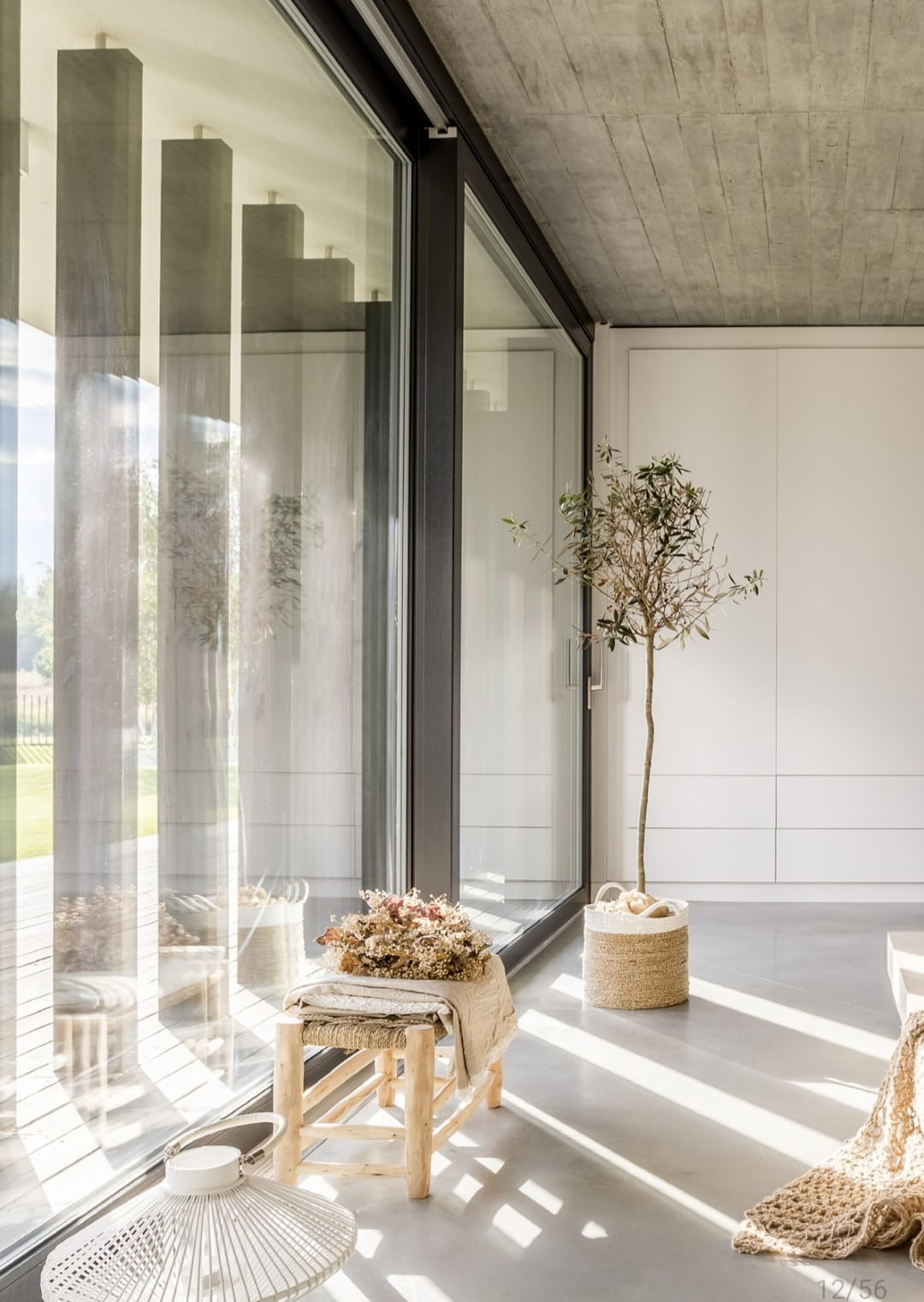 Przesuwne okno tarasowe z czarną ramą w nowoczesnym wnętrzu z betonowym sufitem, oświetlone promieniami słońca padającymi na podłogę i drewniany stołek z dekoracją z suszonych kwiatów.