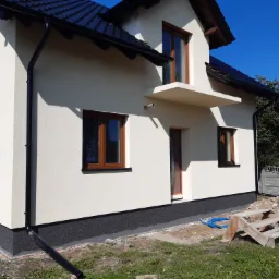 Wykończona elewacja domu jednorodzinnego z ciemną podmurówką i orynnowaniem, widoczne drewniane okna i fragment schodów oraz gruzu.