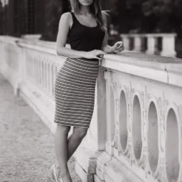 Czarno-białe zdjęcie kobiety opierającej się o kamienną balustradę, ubrana w czarny top i spódnicę w paski, z zamyślonym wyrazem twarzy, w tle widoczny budynek z arkadami.
