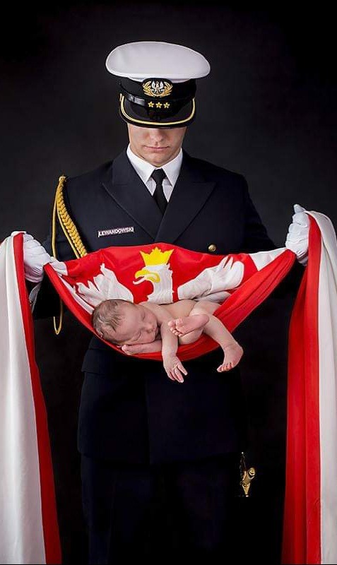Żołnierz w galowym mundurze trzymający w rękawicach niemowlę zawinięte w flagę Polski na czarnym tle.