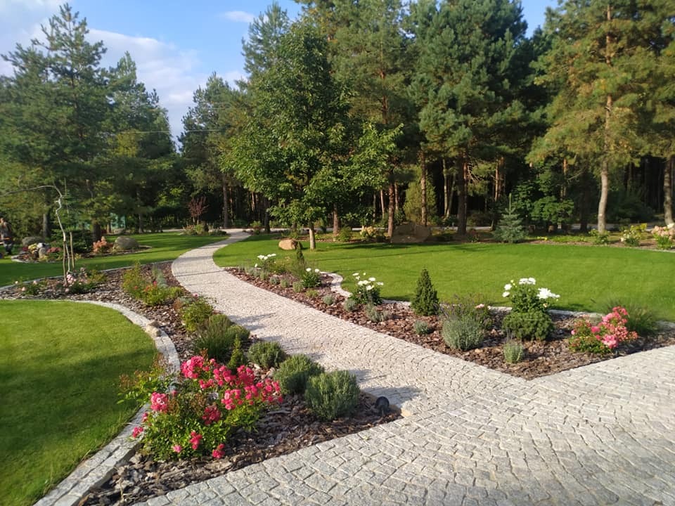 Kręta ścieżka z kostki brukowej prowadząca przez zadbany ogród z równo przystrzyżonym trawnikiem, rabatami kwiatowymi i wysokimi drzewami iglastymi w tle.