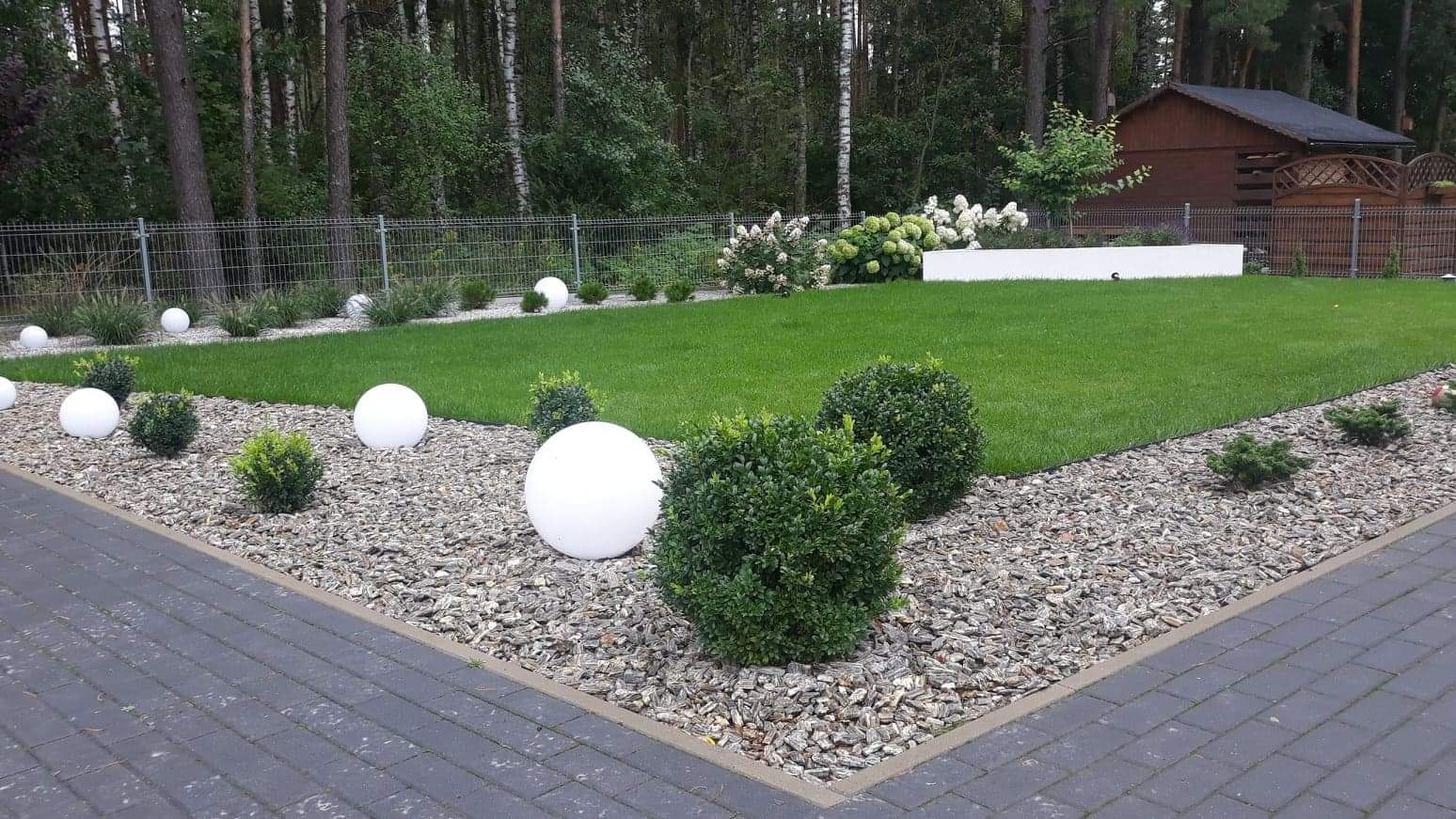 Utrzymany trawnik z kulistymi lampami i krzewami na korze, obok brukowany chodnik, w tle ogrodzenie i drzewa.