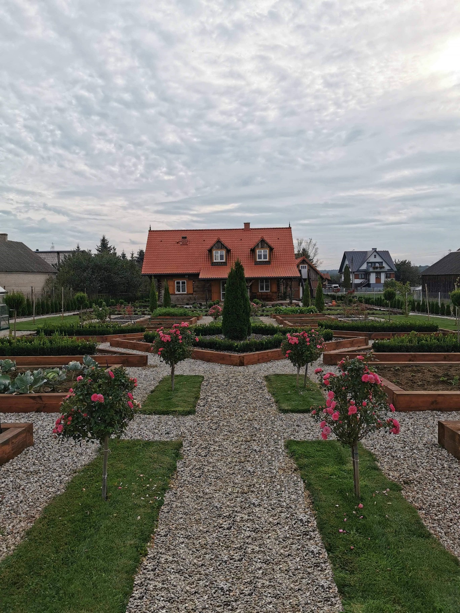 Symetryczny ogród przydomowy z drewnianymi rabatami, ścieżką z drobnego żwiru i różowymi drzewkami różanymi na trawnikach prowadzący do drewnianego domu z czerwoną dachówką.
