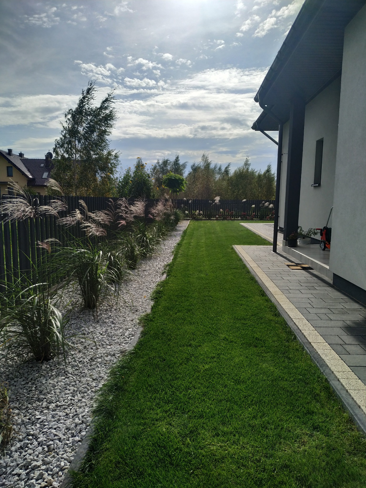 Nowoczesny ogród przydomowy z równo przystrzyżonym trawnikiem, rabatą wysypaną białym grysem i ozdobnymi trawami wzdłuż ciemnego ogrodzenia panelowego.