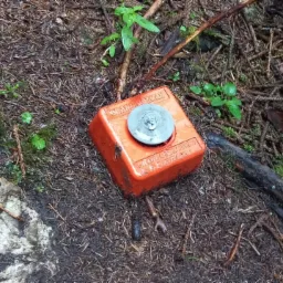 Pomarańczowy, kwadratowy znak geodezyjny z metalową nakładką umieszczony na leśnym podłożu z igliwia, mchu i gałązek.