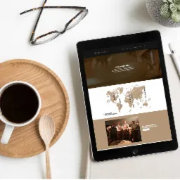 Tablet wyświetlający stronę internetową o tematyce kawowej, obok filiżanka kawy, drewniana łyżka, okulary i roślina w doniczce, wszystko na białym tle.