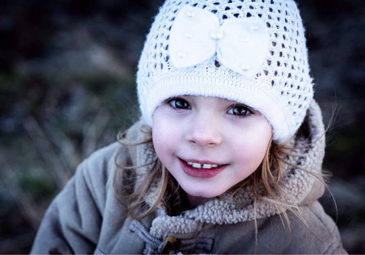 Uśmiechnięta dziewczynka w ażurowej czapce z kokardą, ubrana w kurtkę z barankiem, patrzy w górę, z widocznymi drobnymi ząbkami.