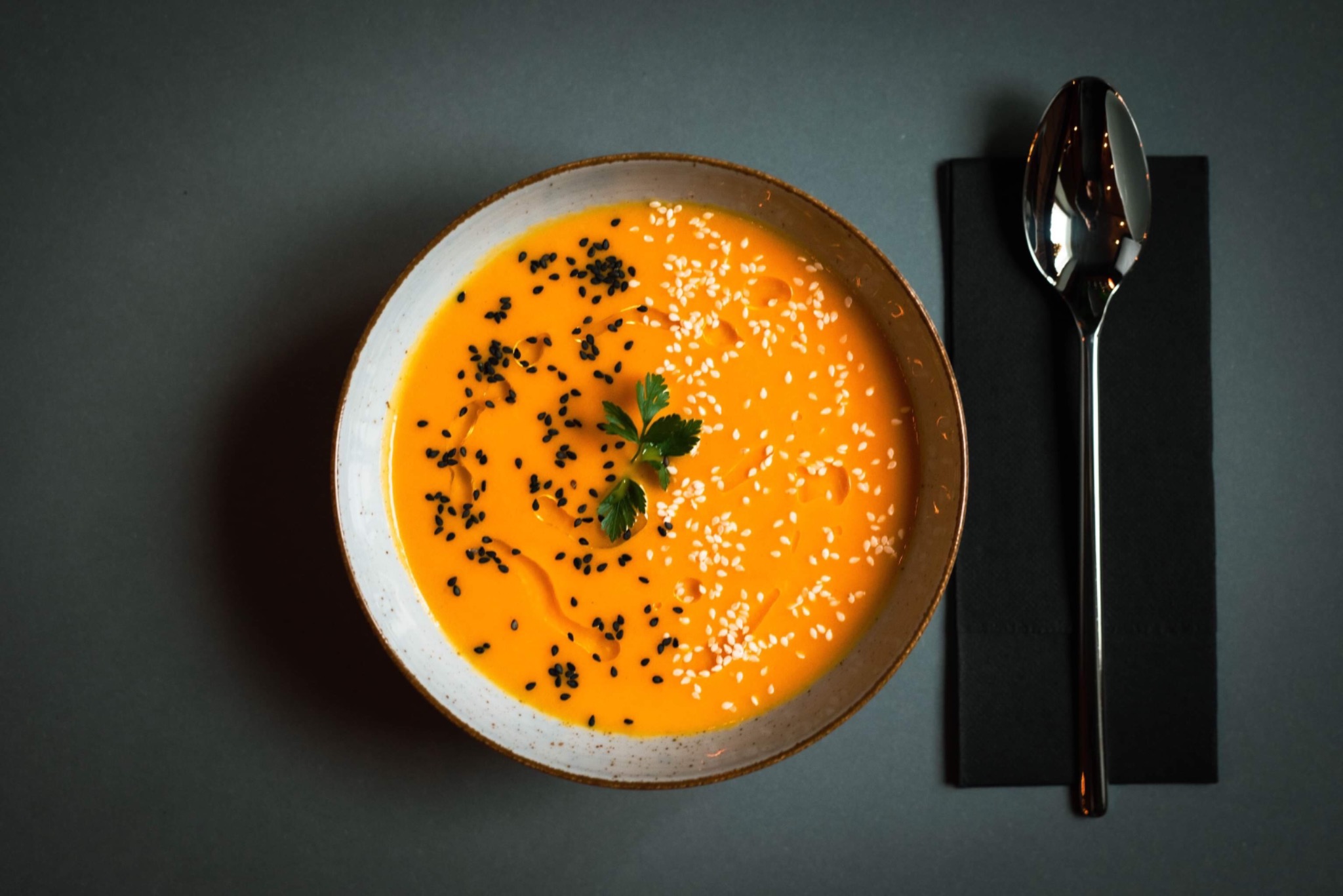 Krem z dyni w ceramicznej misce, posypany czarnym i białym sezamem, udekorowany gałązką pietruszki, obok łyżka na czarnej serwetce, całość na szarym tle.