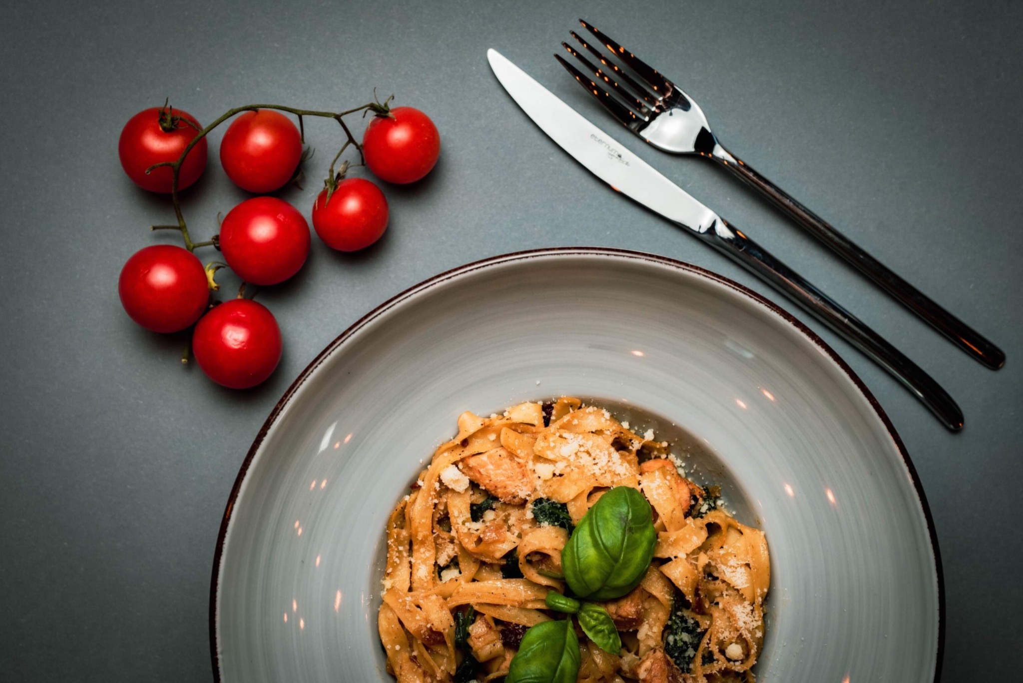 Elegancko podane tagliatelle z kurczakiem, szpinakiem i parmezanem na szarym talerzu, obok gałązka pomidorków cherry oraz sztućce.