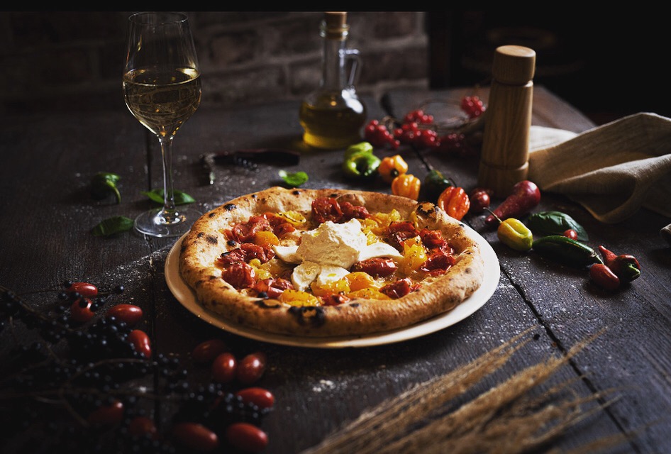 Apetyczna pizza z pomidorami i serem burrata na talerzu, w towarzystwie kieliszka białego wina, oliwy i papryczek chili, na ciemnym drewnianym stole.