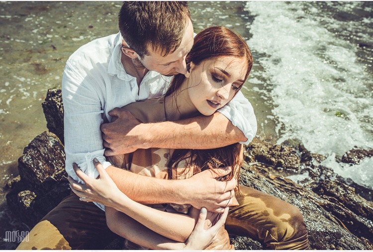 Zakochana para na skalistym brzegu morza, mężczyzna obejmuje kobietę, w tle woda i fale.