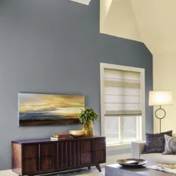 Pokój po remoncie z szarą ścianą, drewnianą komodą z obrazem, beżowymi roletami rzymskimi w oknach, lampą podłogową i narożnikiem w jasnym kolorze.