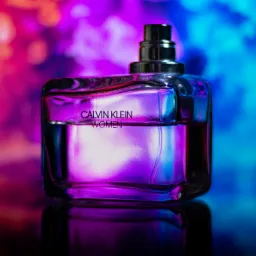 Butelka perfum Calvin Klein Women na czarnym blacie odbijającym światło, z rozmytym tłem w odcieniach różu i błękitu.