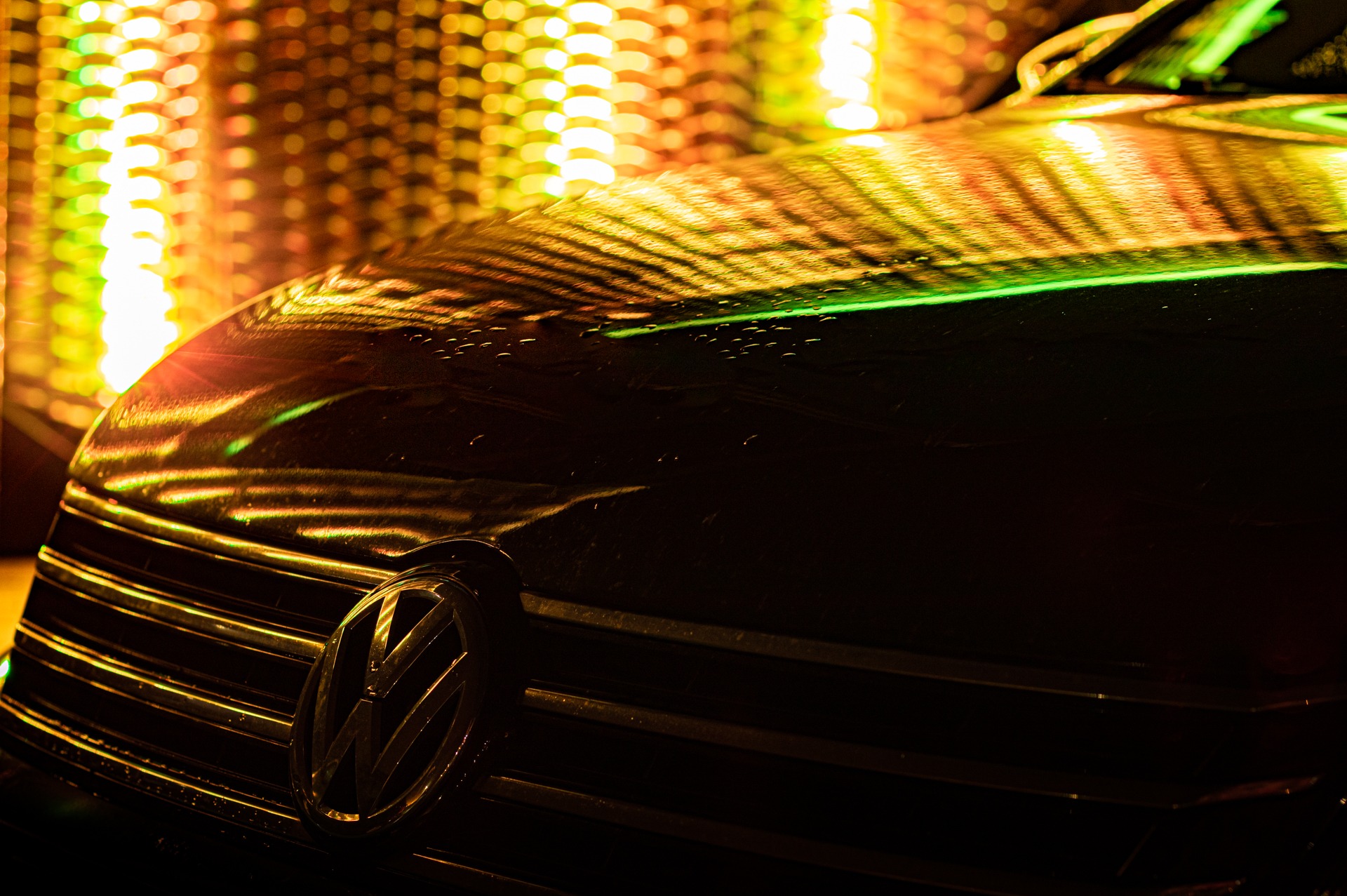 Czarny samochód Volkswagen z chromowanym grillem i logo, oświetlony jaskrawymi żółto-zielonymi światłami, tworzącymi abstrakcyjne wzory na karoserii.