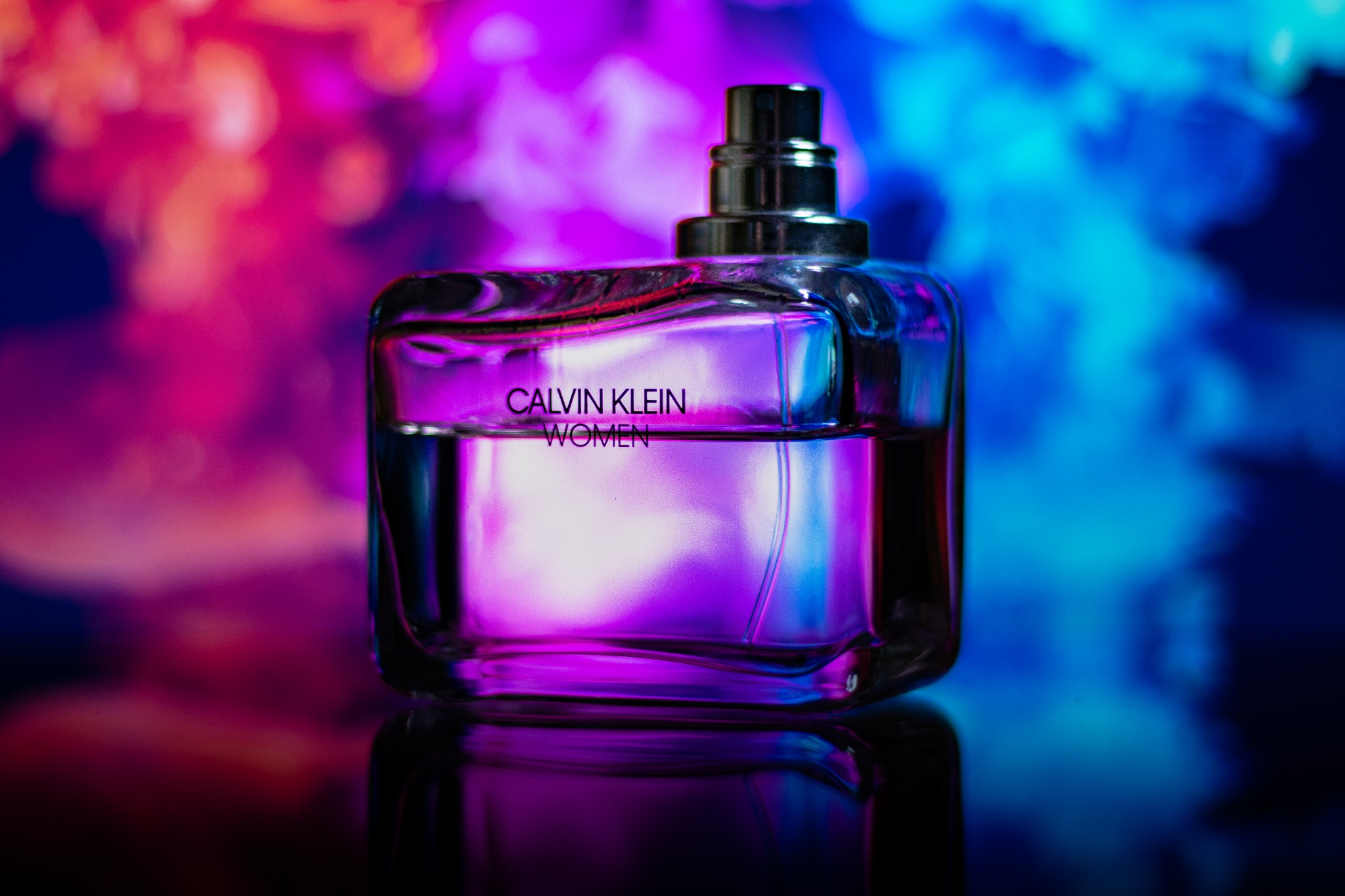 Butelka perfum Calvin Klein Women na czarnym blacie odbijającym światło, z rozmytym tłem w odcieniach różu i błękitu.