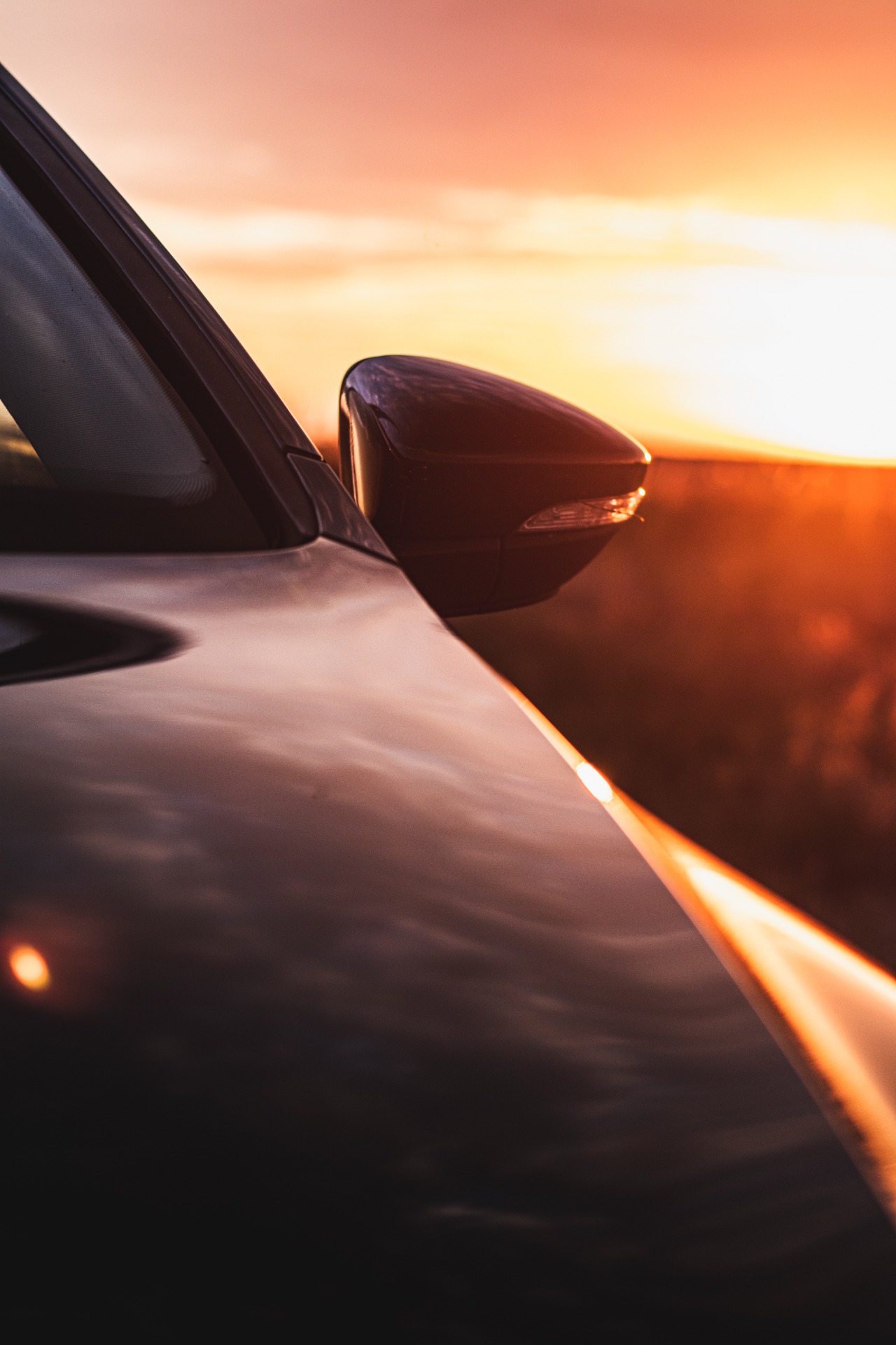 Fragment karoserii samochodu z lusterkiem bocznym na tle zachodzącego słońca, odbijającego się w lakierze.