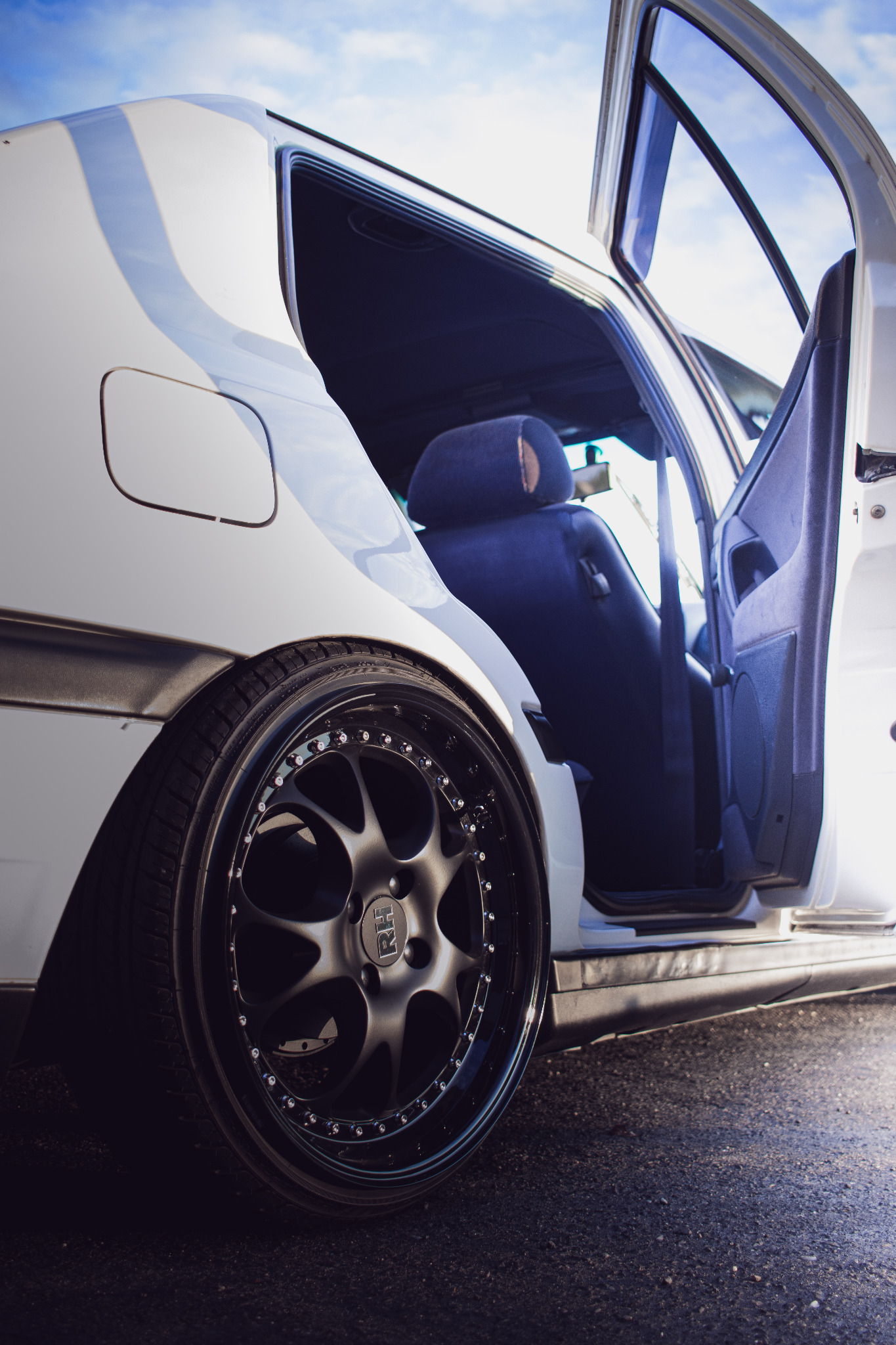 Biały samochód z otwartymi drzwiami, widoczny czarny felgi z logo RH, wnętrze z niebieską tapicerką w tle.