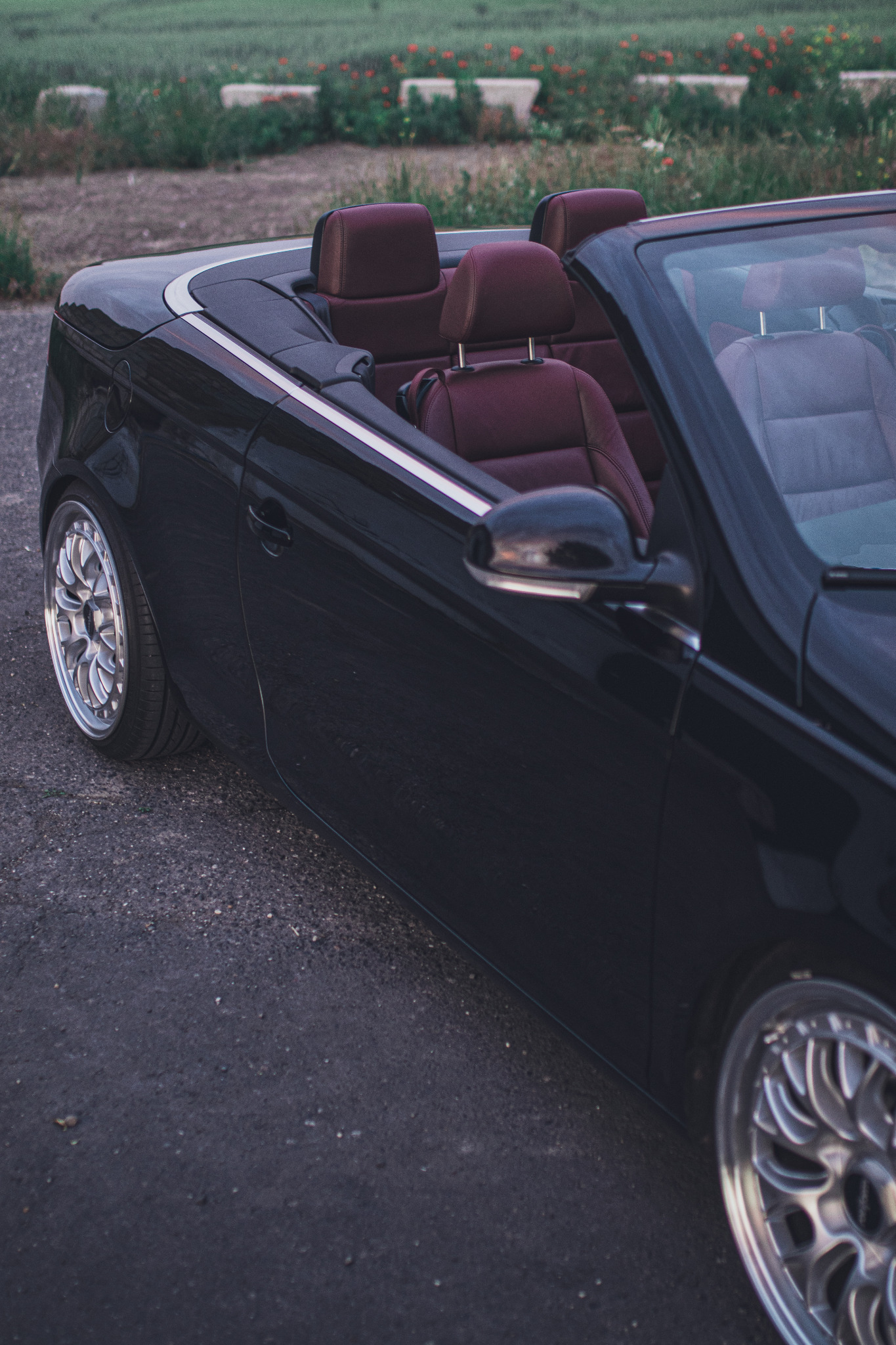Częściowy widok czarnego kabrioletu z burgundowymi skórzanymi siedzeniami i aluminiowymi felgami na asfalcie, z zielonym tłem roślinności.
