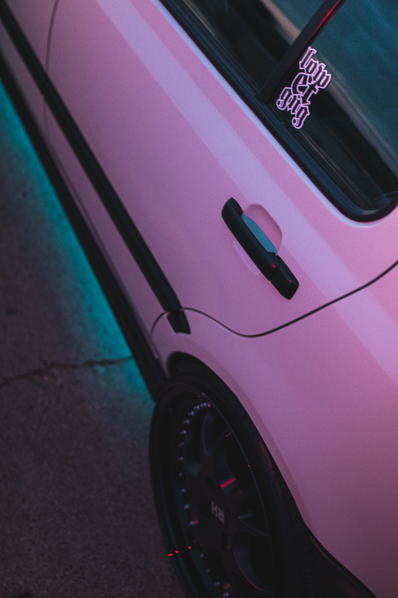 Różowy samochód z czarnymi felgami i naklejką na szybie, oświetlony niebieskim światłem od dołu.
