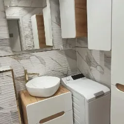 Nowoczesna łazienka z umywalką nablatową na szafce z drewnianym blatem i białymi frontami, pralka automatyczna obok, marmurowe płytki na ścianach, złota armatura i rama lustra.