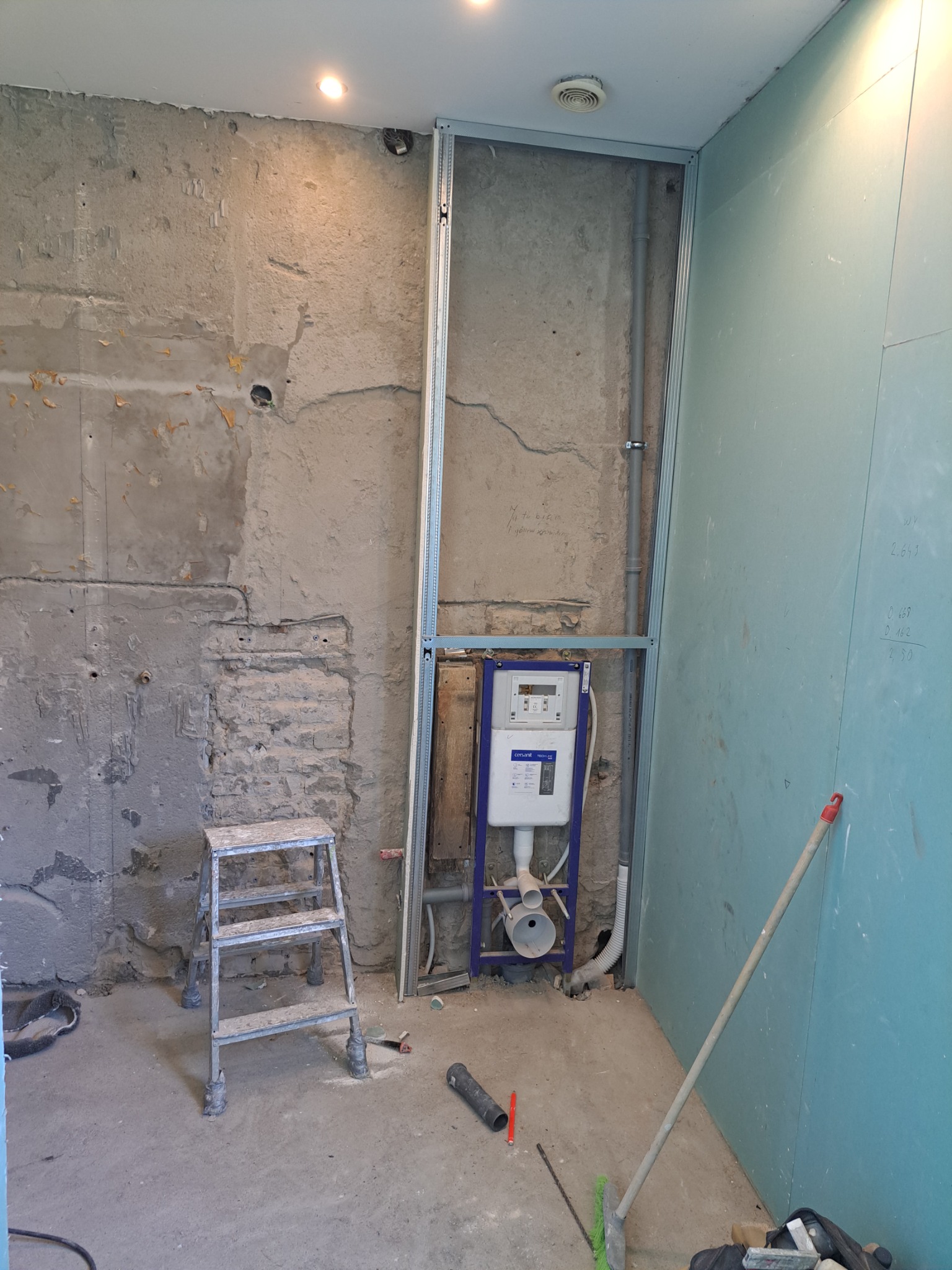 Stelaż podtynkowy WC zamontowany w metalowej konstrukcji przy surowej ścianie, obok płyta gipsowo-kartonowa, drabinka i narzędzia na podłodze.