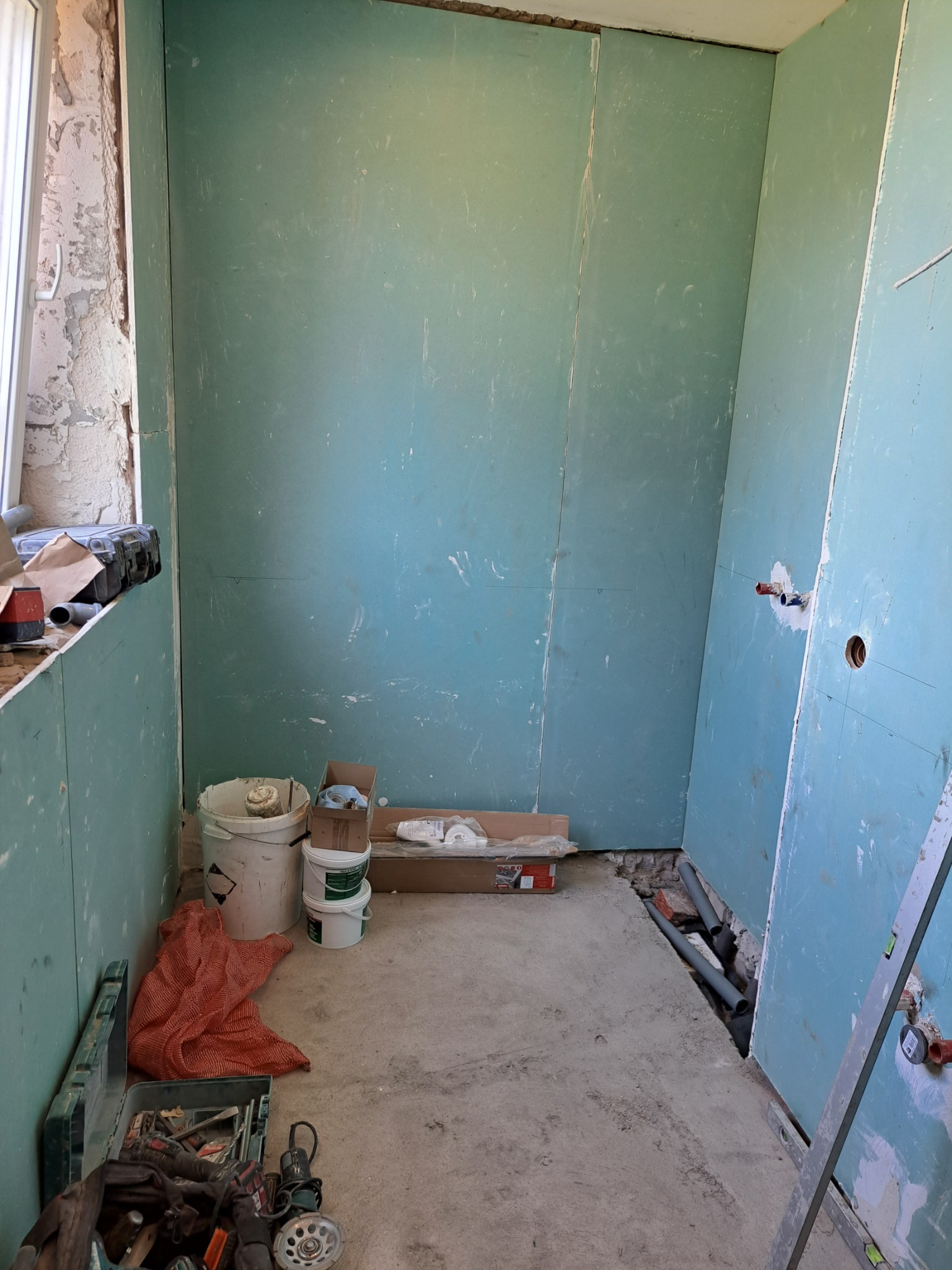 Wnętrze łazienki w trakcie prac przygotowawczych: ściany obłożone płytami gipsowo-kartonowymi, widoczne instalacje wodne, narzędzia i materiały budowlane w nieładzie.