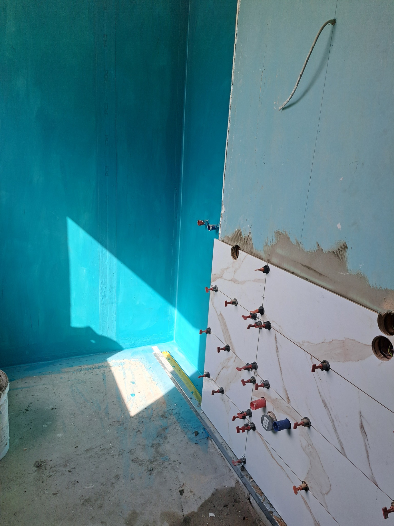 Niedokończona łazienka z hydroizolacją w turkusowym kolorze, białe płytki imitujące marmur mocowane za pomocą systemu poziomowania płytek.