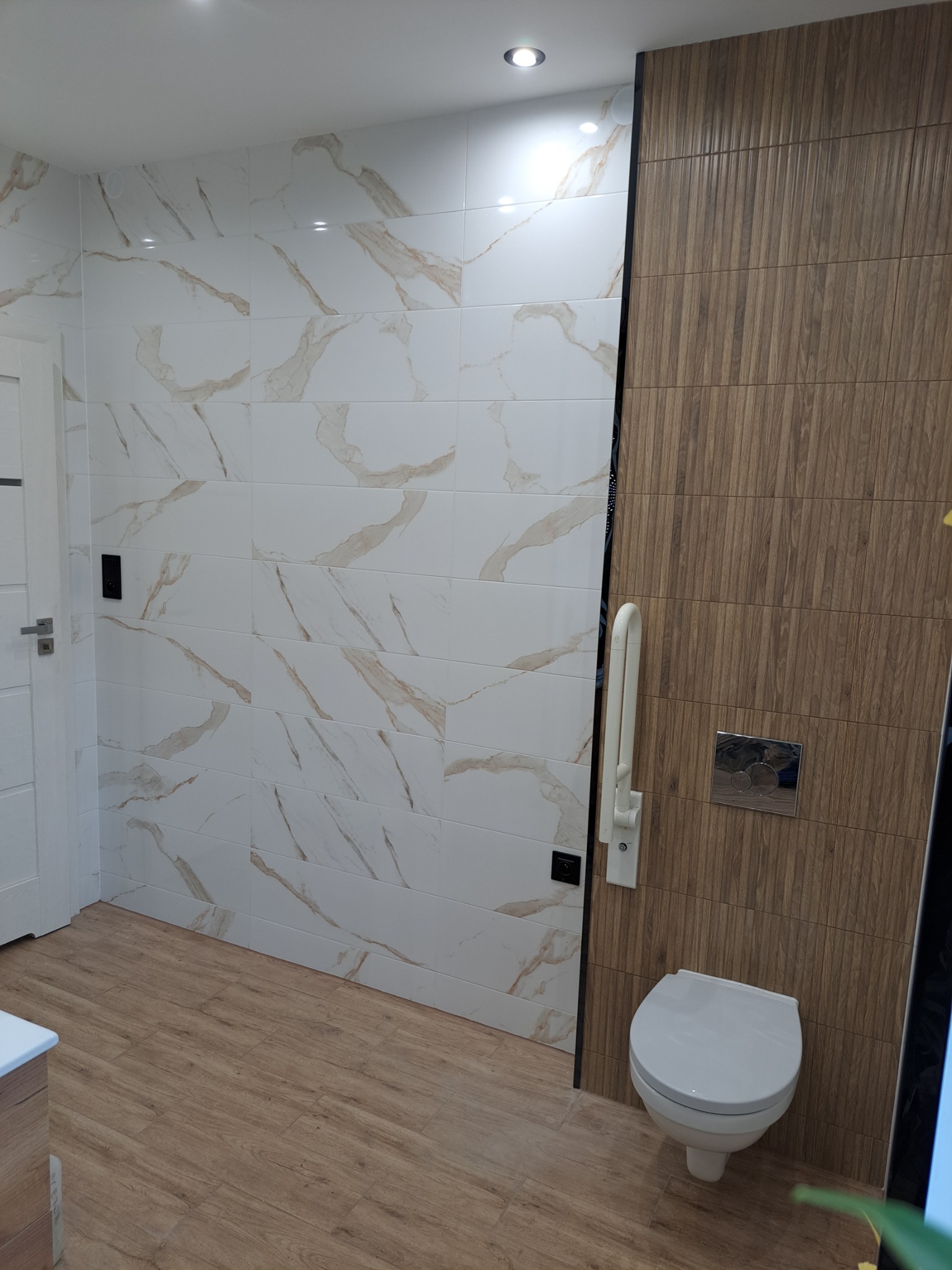 Nowoczesna łazienka z marmurowymi płytkami na ścianie, drewnopodobnymi płytkami na drugiej ścianie, wiszącą toaletą z chromowanym przyciskiem i drewnopodobną podłogą.