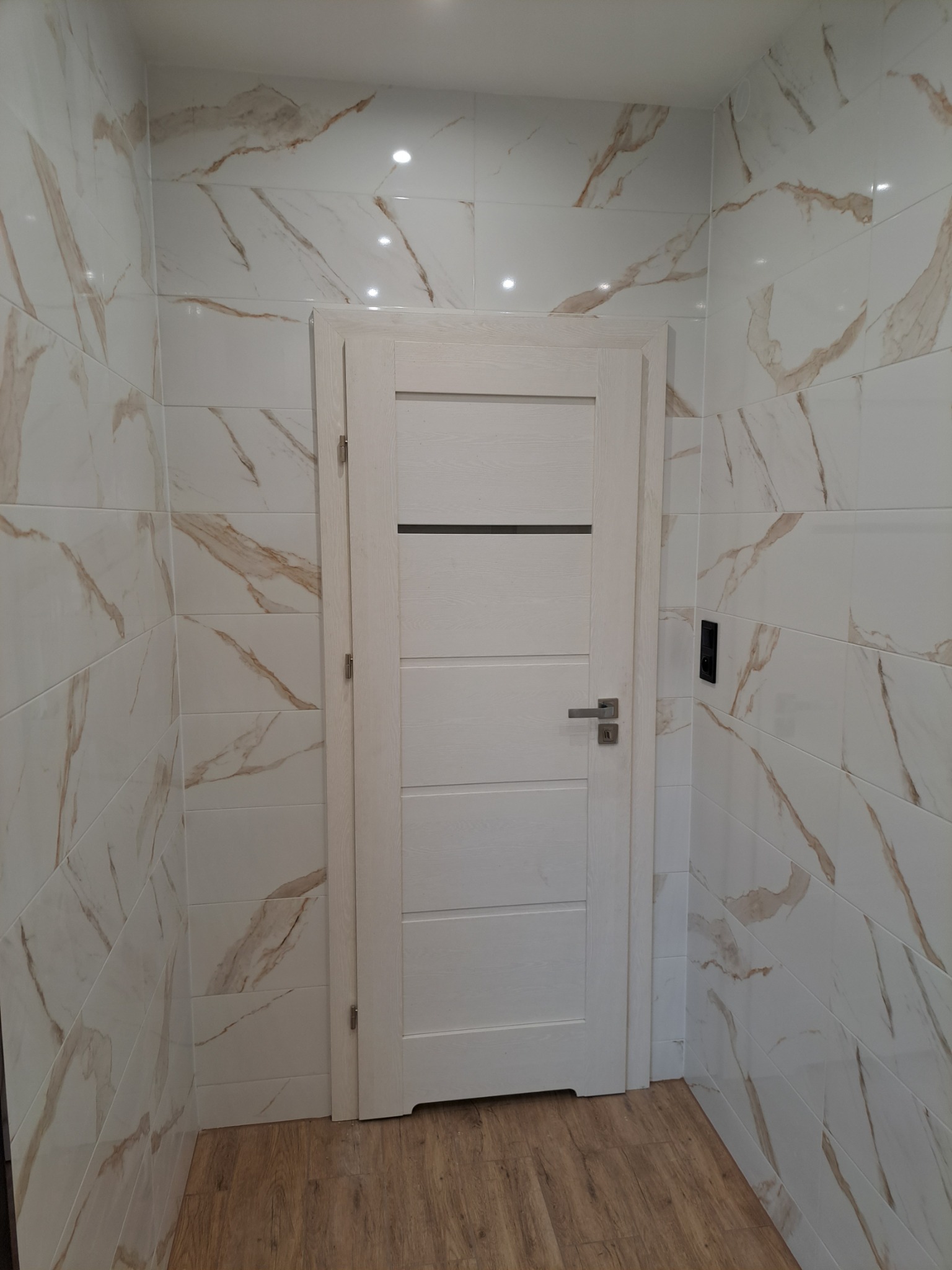 Wykończona łazienka z biało-złotymi płytkami imitującymi marmur, drewnopodobna podłoga i białe drzwi z poziomymi frezowaniami oraz czarnym akcentem.