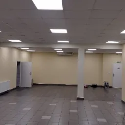 Przestronne wnętrze komercyjne po odświeżeniu ścian, z widoczną drabiną i urządzeniem malarskim, sugerującym prace wykończeniowe.