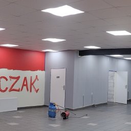 Bud-Walczak Mariusz Walczak - Wnętrze biura w trakcie malowania: część ściany pomalowana na czerwono z napisem 'Bud-WALCZAK', reszta ścian szara, widoczny grzejnik, drabina, nagrzewnica gazowa, oraz pracownik z wiadrem farby.