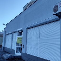 Bud-Walczak Mariusz Walczak - Szary budynek usługowy z zamkniętymi roletami antywłamaniowymi, klimatyzator LG na elewacji i ogłoszeniem 'Sprzedam/Wynajmę' w drzwiach wejściowych.