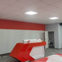 Bud-Walczak Mariusz Walczak - Wnętrze biura z pomalowanymi na czerwono ścianami i białymi panelami ściennymi, widoczna recepcja z opakowaniem folii Oracal, sufit z kwadratowymi lampami.