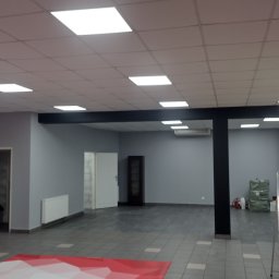 Bud-Walczak Mariusz Walczak - Przestronne wnętrze biurowe w trakcie odświeżania, z elementami czerwieni i szarości, widoczna drabina i przygotowane materiały budowlane, jasne oświetlenie sufitowe.