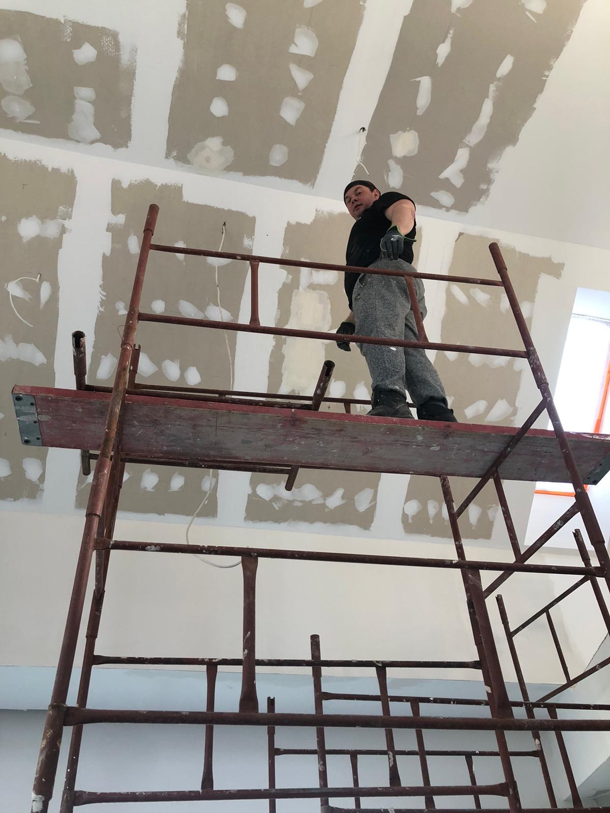 Pracownik na rusztowaniu, szpachluje łączenia płyt karton-gips na suficie, widoczne ślady szpachli i świeżo pomalowane fragmenty sufitu.