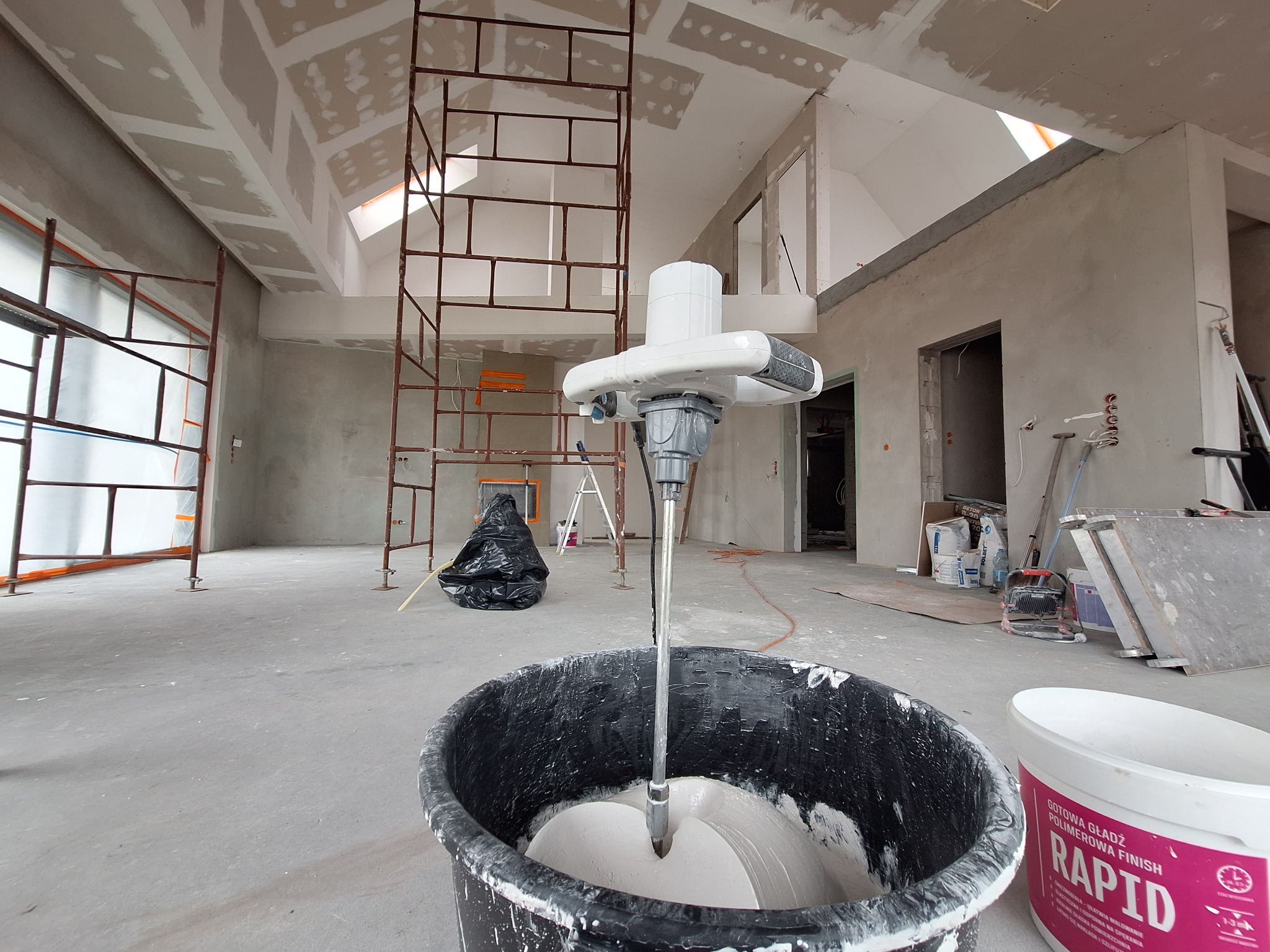 Mieszanie gotowej gładzi polimerowej Rapid w czarnym wiadrze za pomocą mieszadła elektrycznego na tle surowego wnętrza z rusztowaniami i płytami kartonowo-gipsowymi na suficie.