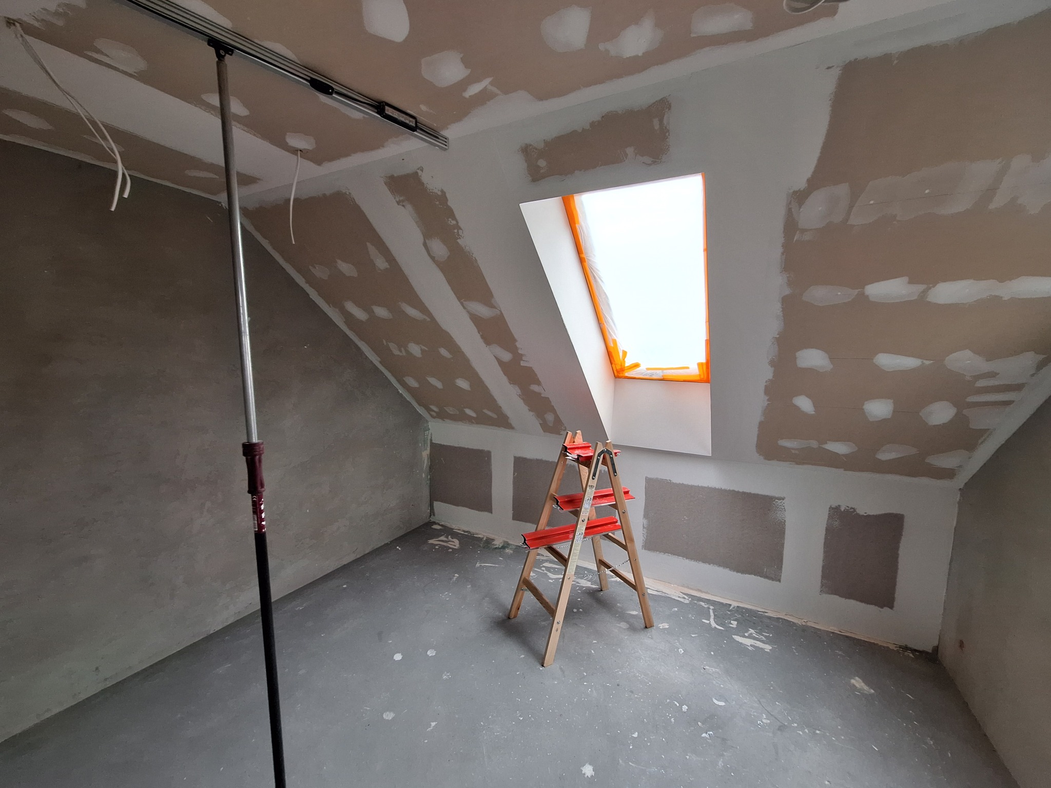 Adaptacja poddasza: ściany i sufit z płyt kartonowo-gipsowych, częściowo zaszpachlowane, okno dachowe zabezpieczone folią, drabina rozstawiona na szarym betonie, widoczny wysięgnik malarski.